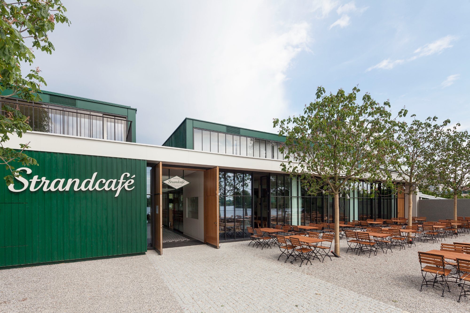 Außenansicht des Strandcafés mit grüner Fassade, offenem Eingang und Terrasse aus Holztischen und Stühlen, Bäume im Umfeld.
