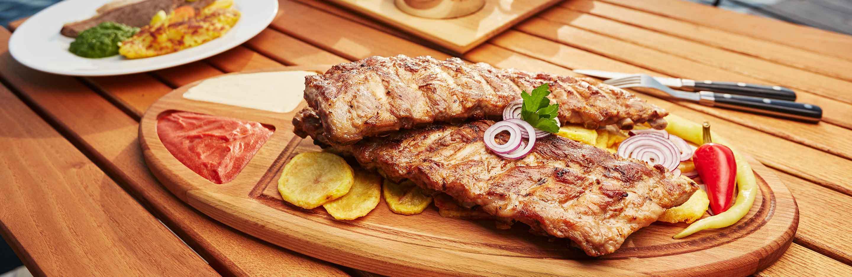 Gegrillte Rindersteaks auf einem Holzservierbrett mit Kartoffelchips, Zwiebelringen, roter Paprika und Petersilie – Outdoor-Tisch.