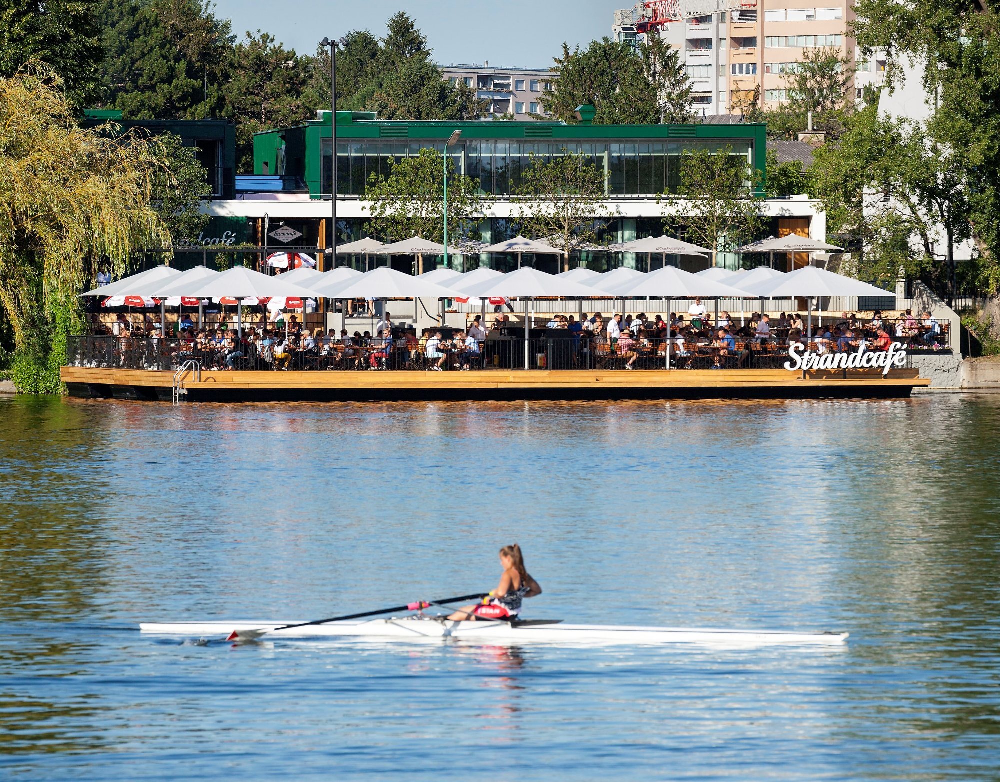 Ruderer in Einmannboot rudert auf dem Fluss; dahinter schwimmendes Strandcafé mit weißen Sonnenschirmen und Gästen.