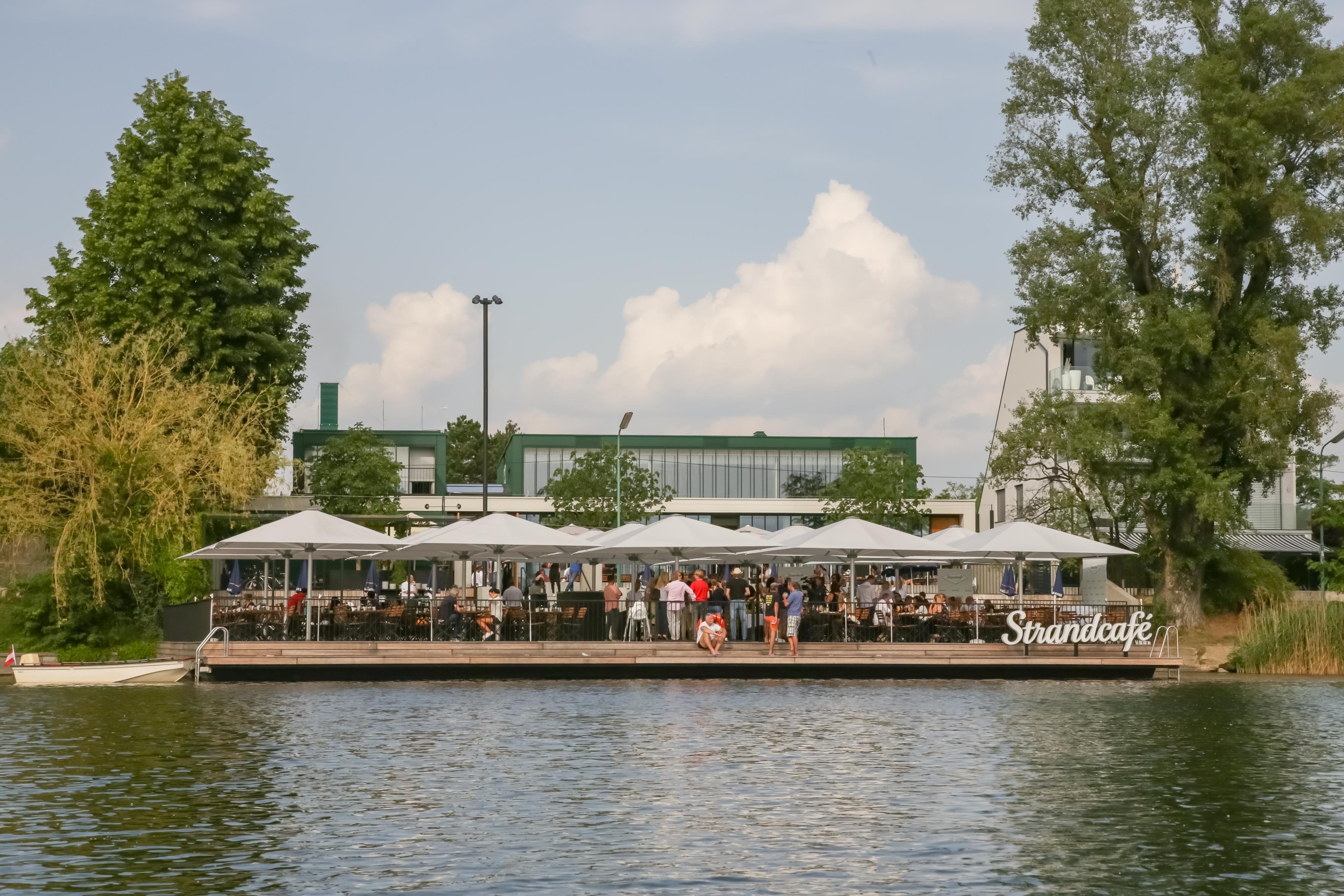 Strandcafé auf Holzdeck am See mit weißen Sonnenschirmen; Gäste speisen und plaudern, dahinter Bäume und Gebäude.
