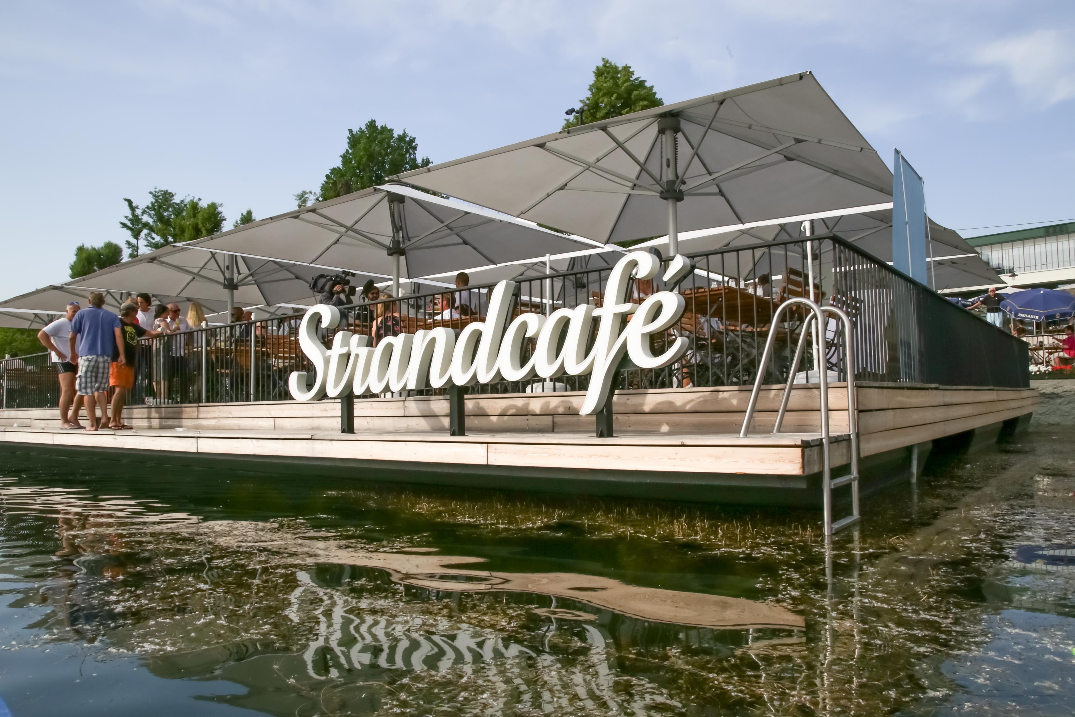 Schwimmendes Strandcafé mit Terrassen unter Sonnenschirmen; Besucher am Deck, sichtbarer Schriftzug "Strandcafe".