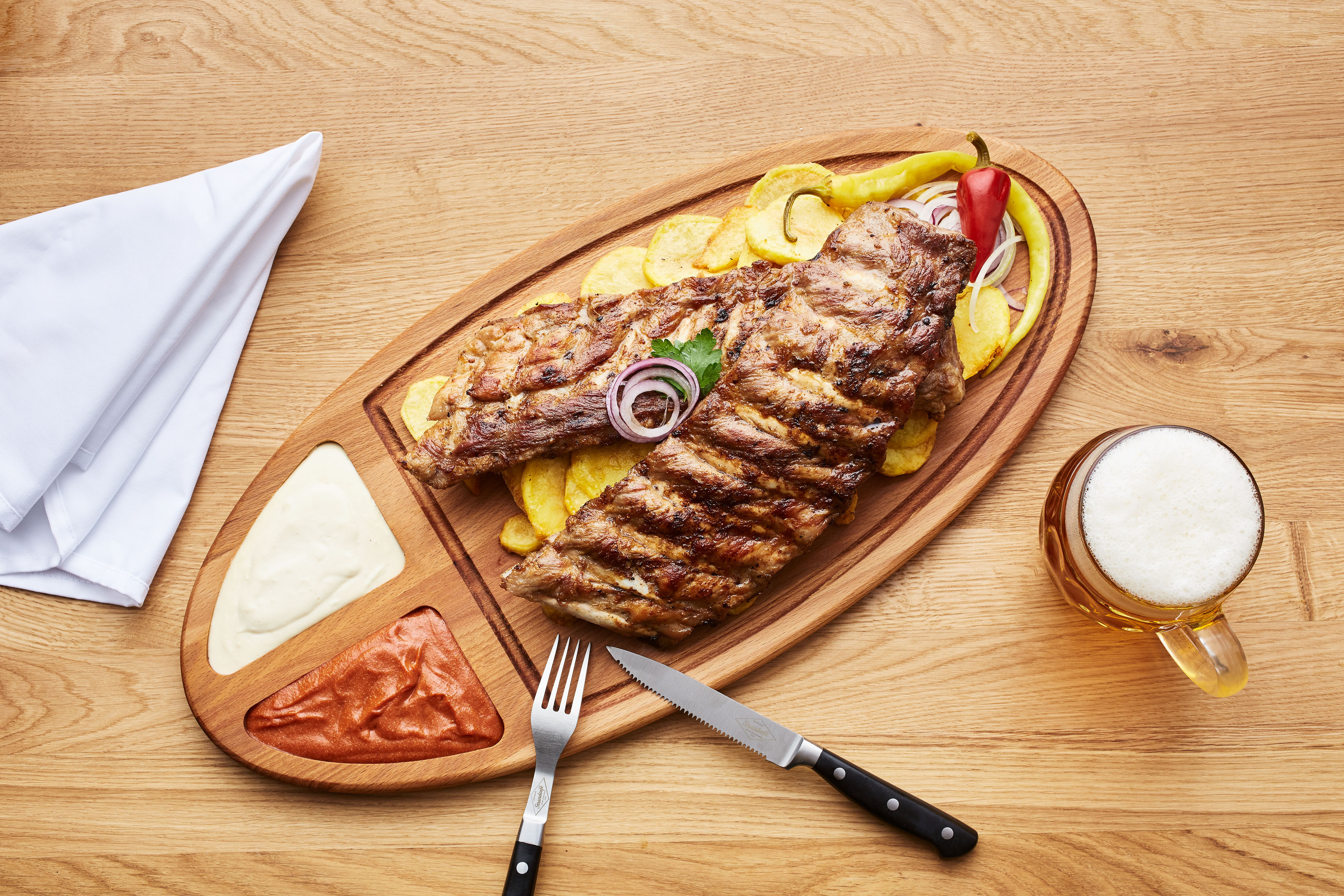 Gegrilltes Rindersteak auf Kartoffel-Chips mit Zwiebelringen; Paprika-Garnitur, Saucen auf Holzbrett, Bierkrug daneben.