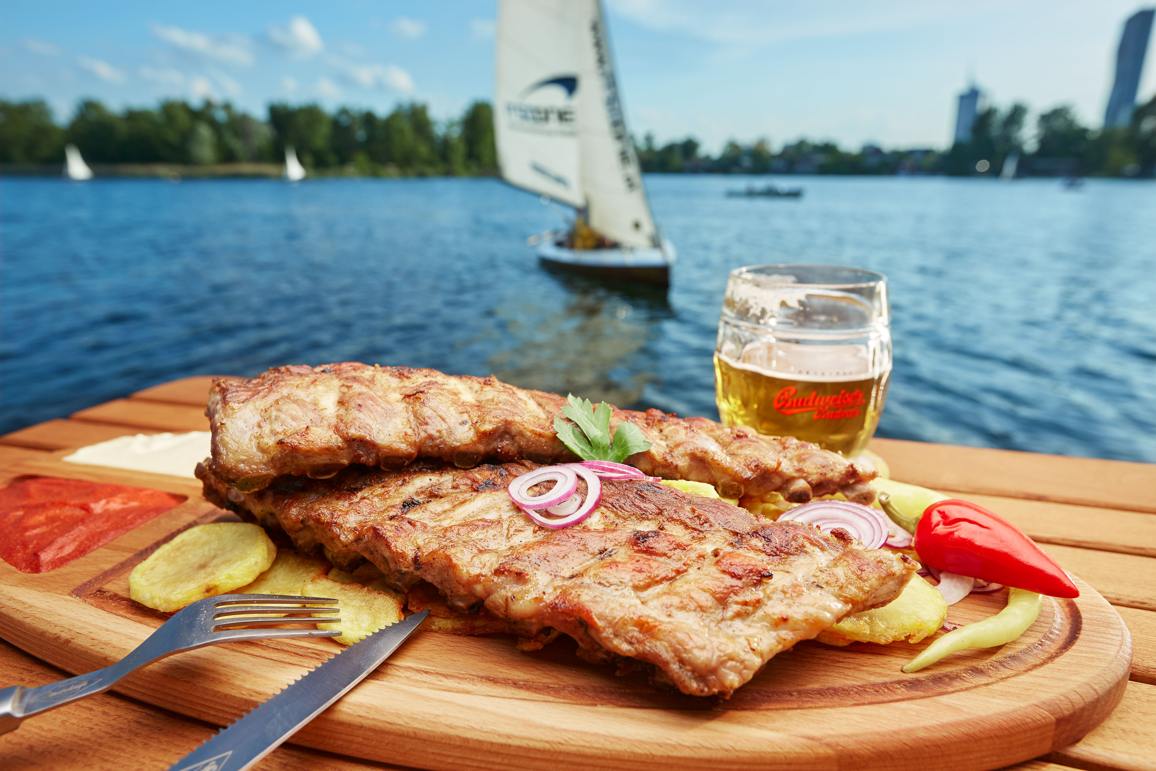 Gegrillte Schweinerippchen auf Holzteller mit Zwiebeln, Kartoffelchips am See; Segelboot im Hintergrund, Bierglas.
