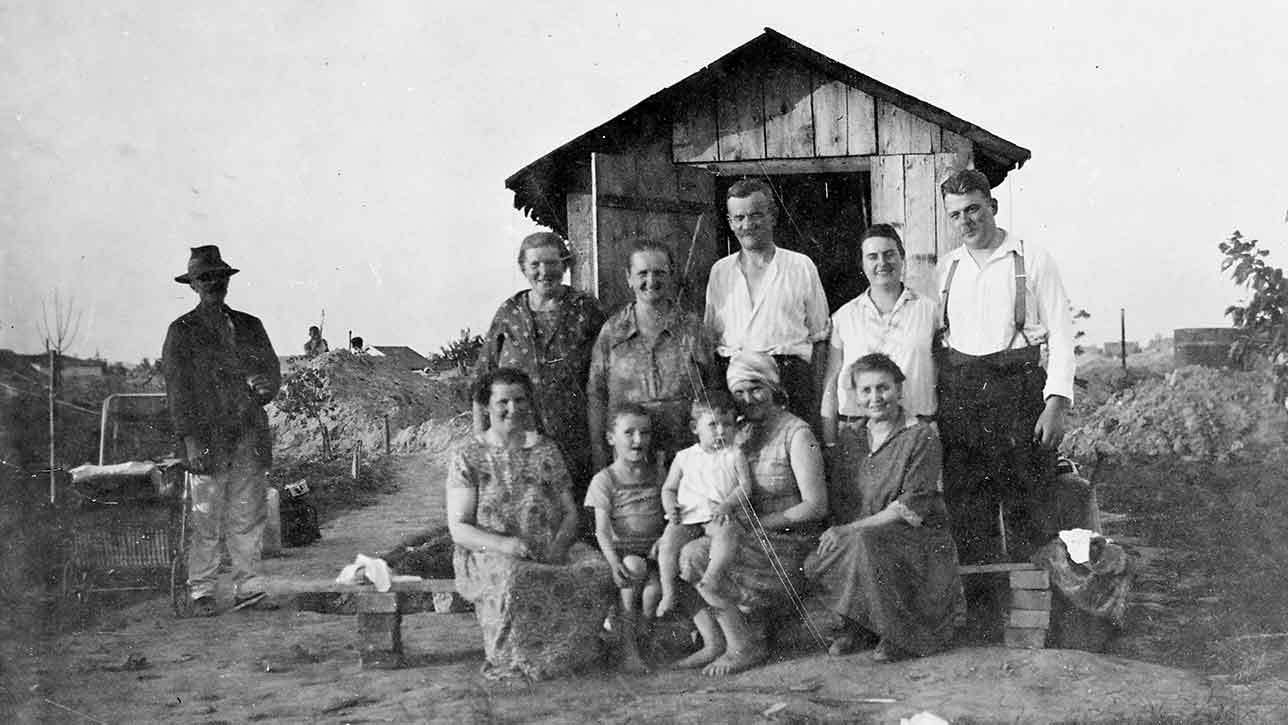 Familiengruppe posiert vor kleinem Holzhäuschen im Freien; mehrere Erwachsene und Kinder stehen und sitzen.