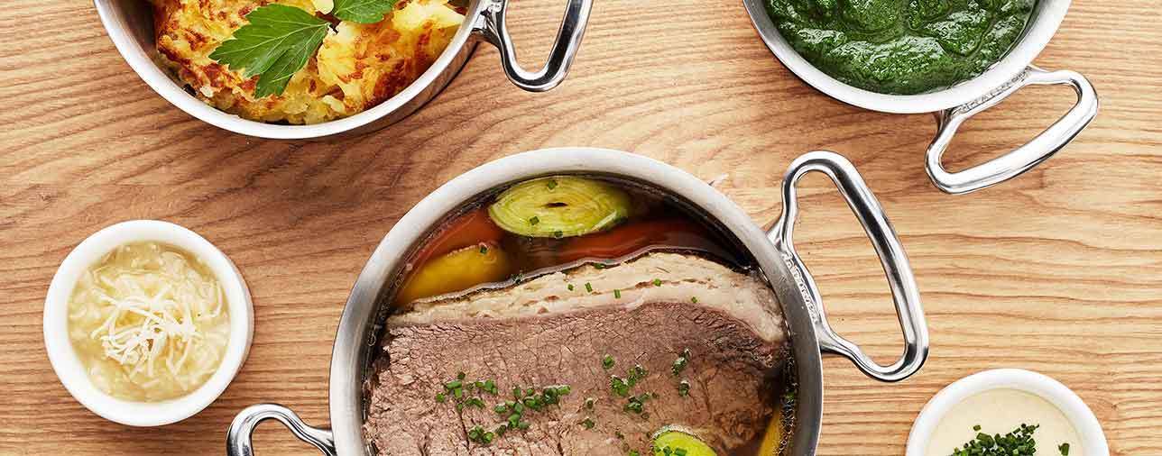 Rinderbraten im Schmortopf in Brühe mit Gemüse, auf Holztisch; daneben Gratin, grüne Sauce und Käse-Dip.